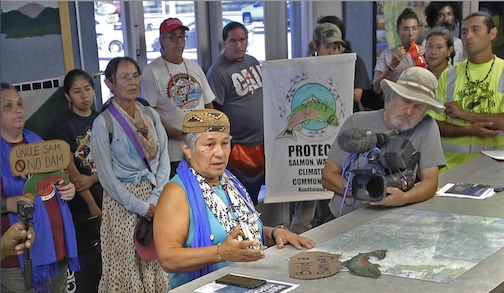 A woman sits at a table, speaking out with a group of allies behind her. A person in a hat films the speaker on the right.