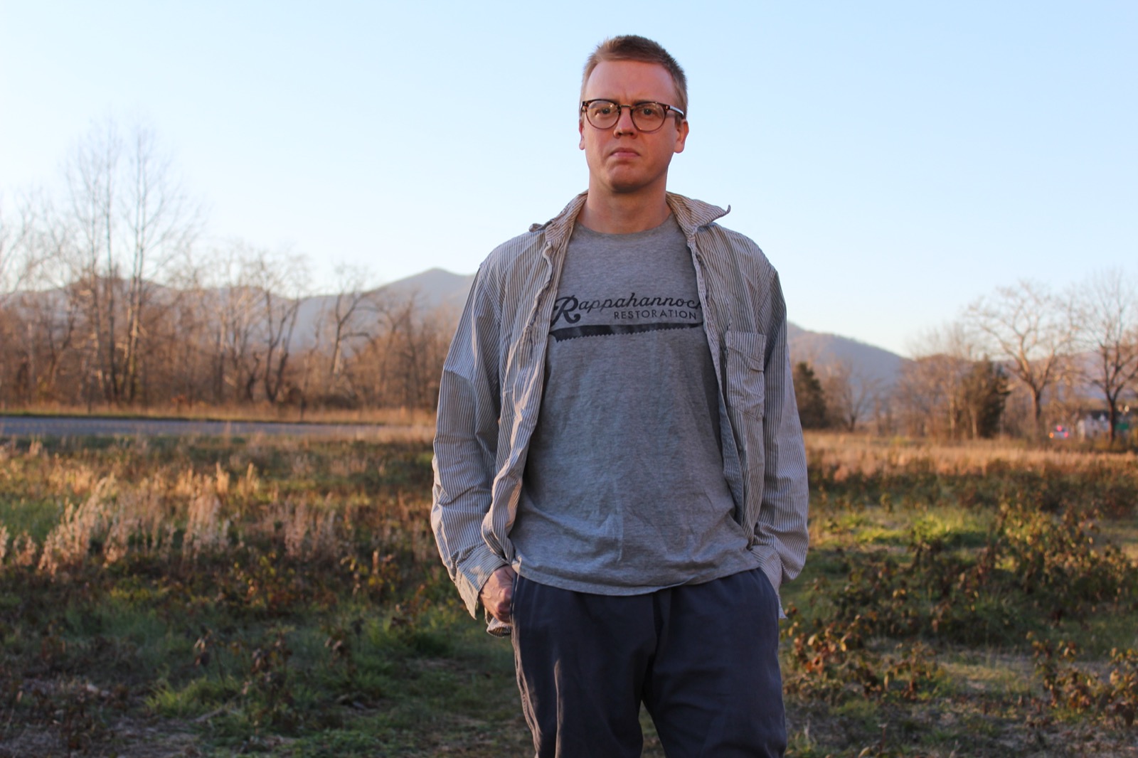 a man wearing a grey open button shirt with a grey tshirt beneath it. behind him is a grass field and a mountain