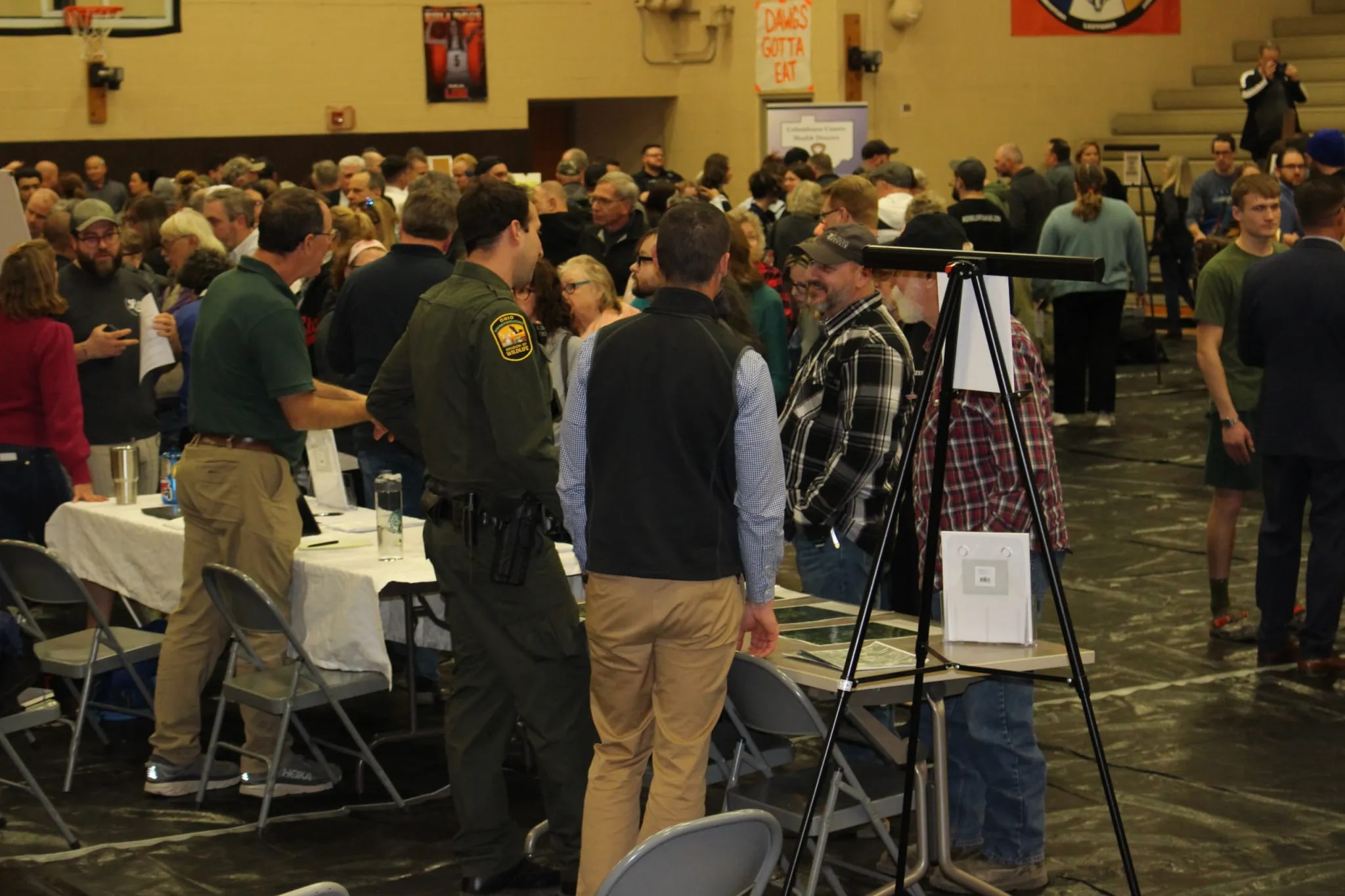 a crowded auditorium, with a few tables set up. a cop and a few other people behind the tables talk to a larger crowd on the other side of the tables