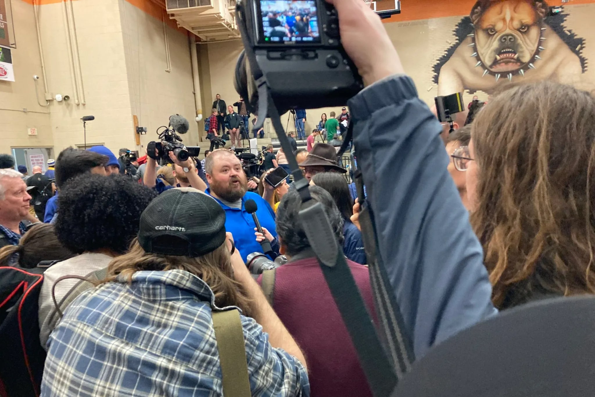 a crowd of reporters gather around a man in a blue shirt, someone is holding up a mic near him