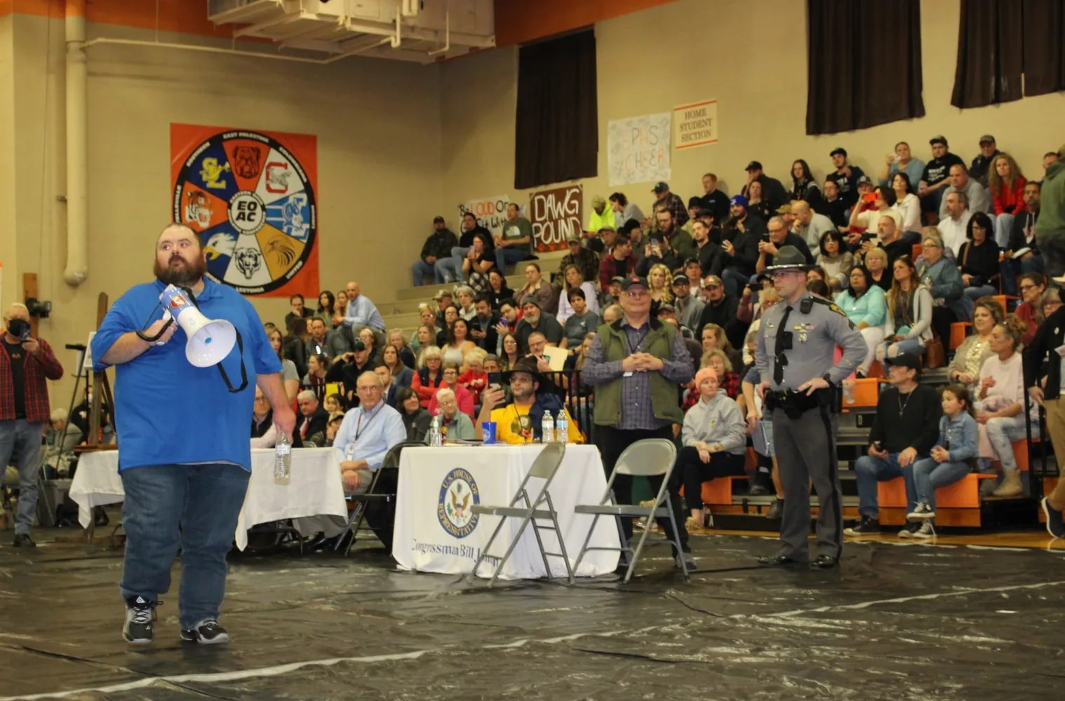 a man holding a megaphone walks in front of a crowd on bleachers and a cop seated in an auditorium