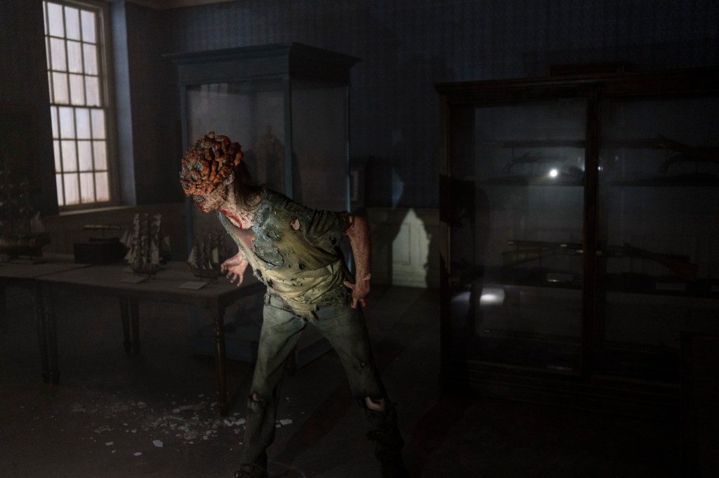 A zombie with blooming fungal outbreak on its face roars inside a dark room.