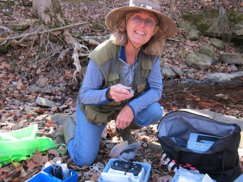 A woman in a wooded area kneels at the ground, picking up small samples of water in a test tube.