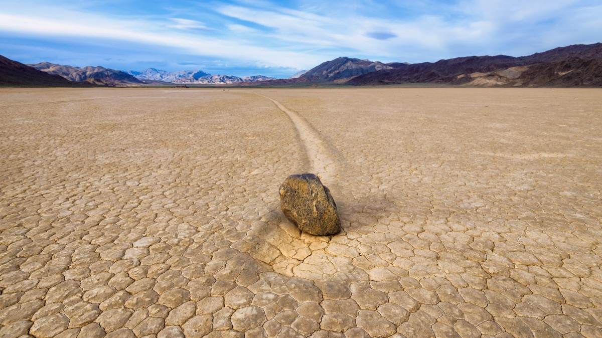 Dry, tan ground that is cracked. In the distance are mountains. In the forground is a roughly shaped rock. The rock sits in a groove cut into the ground.