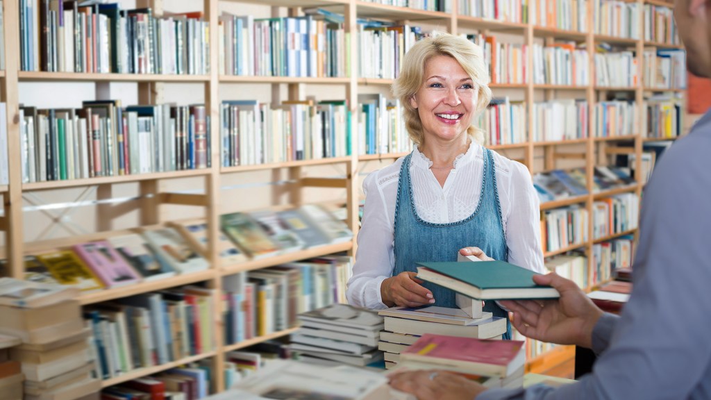 Smiling woman taking book in bookstore, talking and handing a book to another person out of frame
