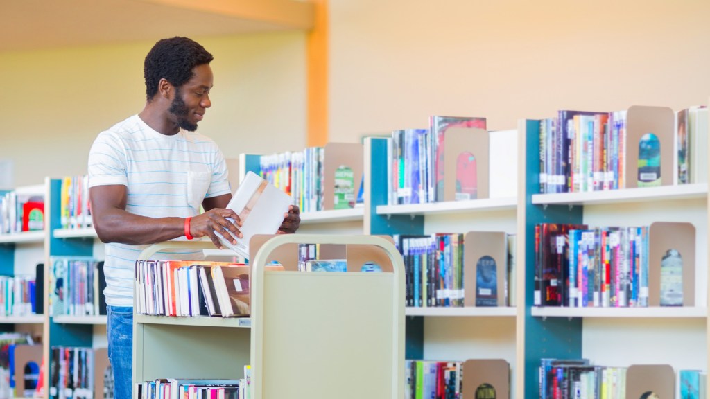 A man at a library cart, holding a book and arranging books in library