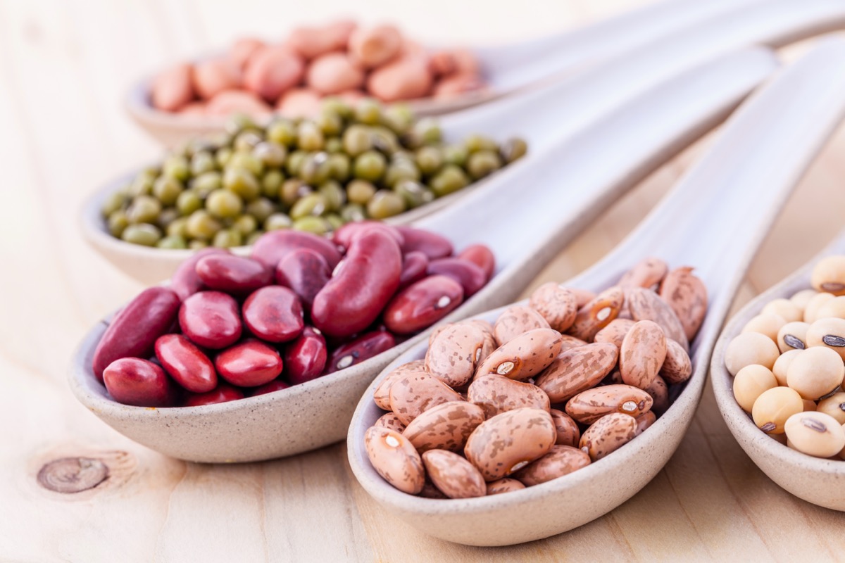 Dried kidney beans, pinto beans, black-eyed peas, and other seeds arranged in white ceramic spoons on a wood table.