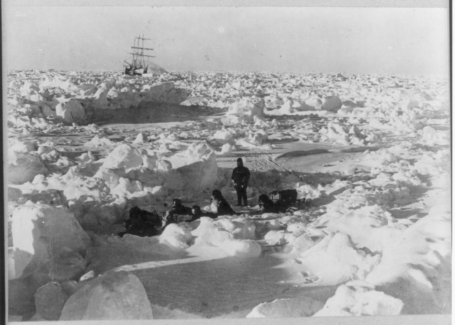 crew member with dog team hitched together sitting among ice floe; in the far background the Endurance ship is stuck.
