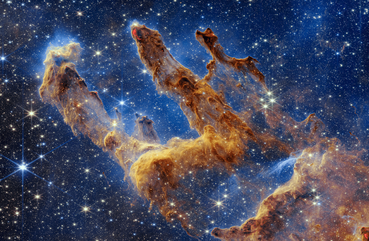 deep in space, orange dense yet wispy pillars look like arches and spires rising out of a desert landscape, but are filled with semi-transparent gas and dust. This is a region where young stars are forming – or have barely burst from their dusty cocoons as they continue to form.