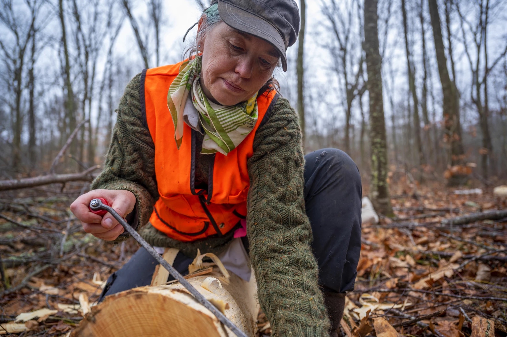 A person in a visibility vest cuts bark off of a log in the forest.