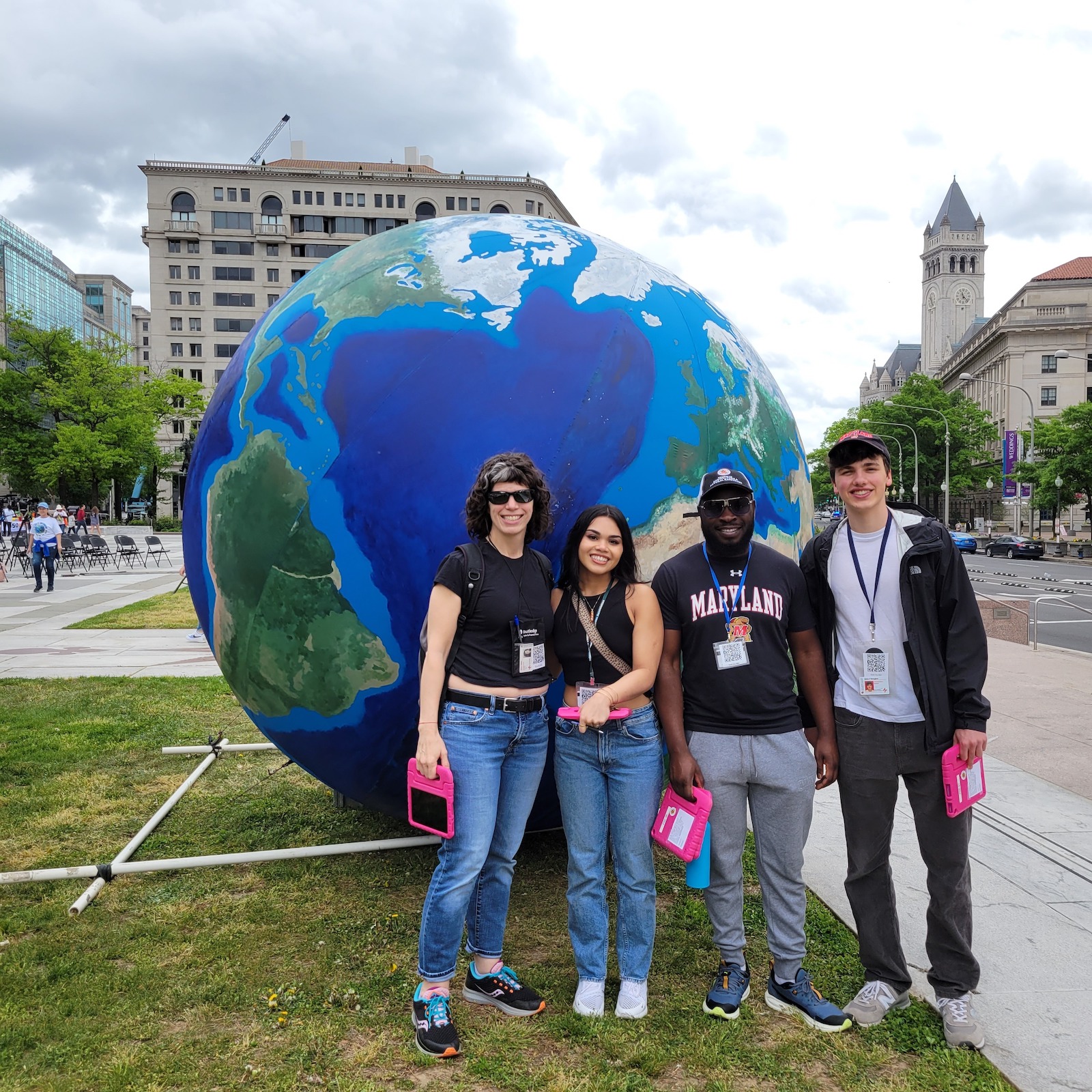 outside on a grassy path next to a street, four people, a female professor and three students, smile and stand in front of a model of the earth, that stands about four or five feet taller than the people.