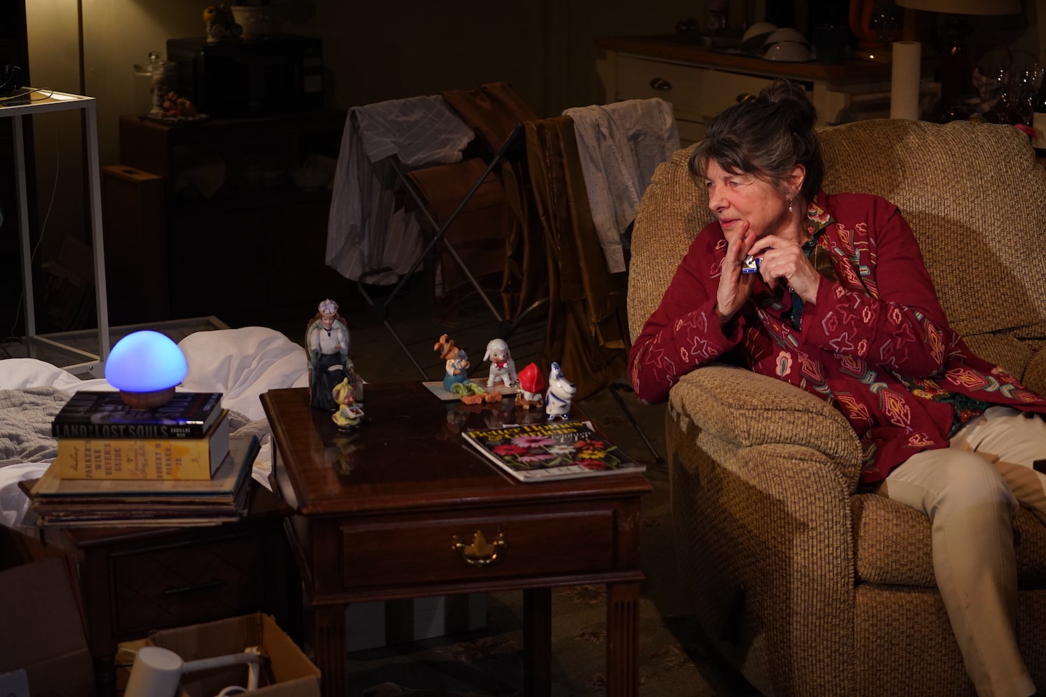 a stage set of a living room. an older woman sits in a brown recliner chair next to a side table of an assortment of animal figurines and knick knacks. further left is a soft blue glowing dome that's a smart speaker