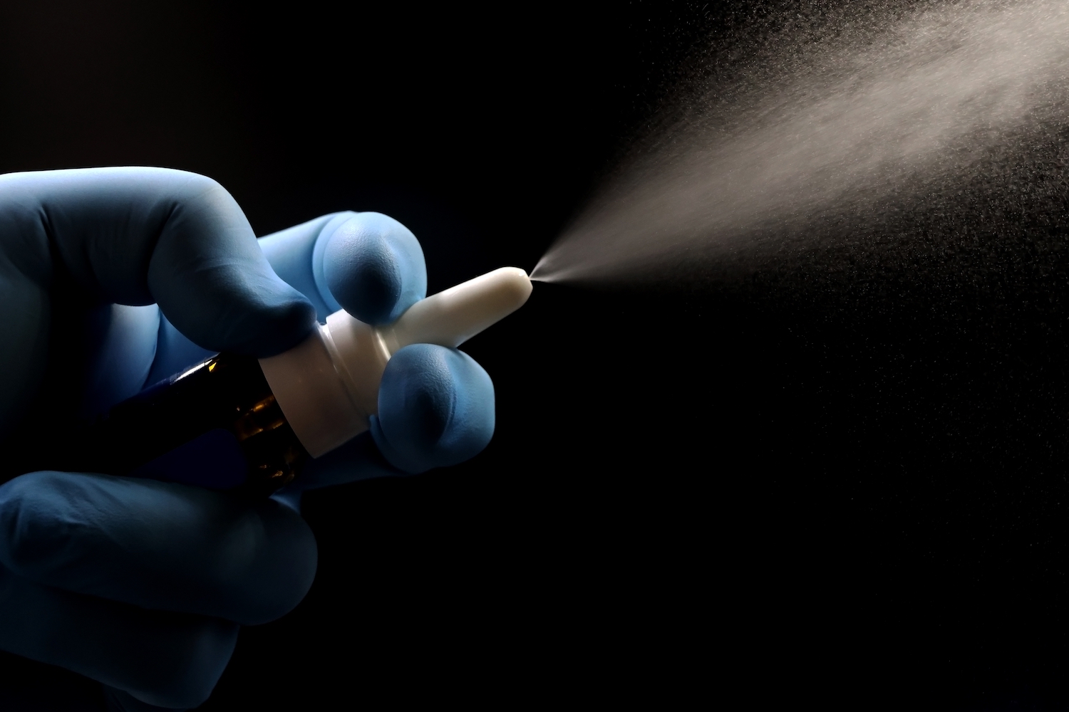 a hand squeezes a bottle of vaccine projecting mist spray, on a black background 