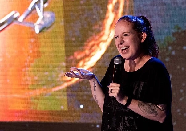 an astrophysicist with really cool tattoos on her arm and with her hair tied in a ponytail smiles and talks into a mic with her hand propped out in gesture. behind her is a screen display of a futuristic space ship, from the star trek series, flying towards a planet