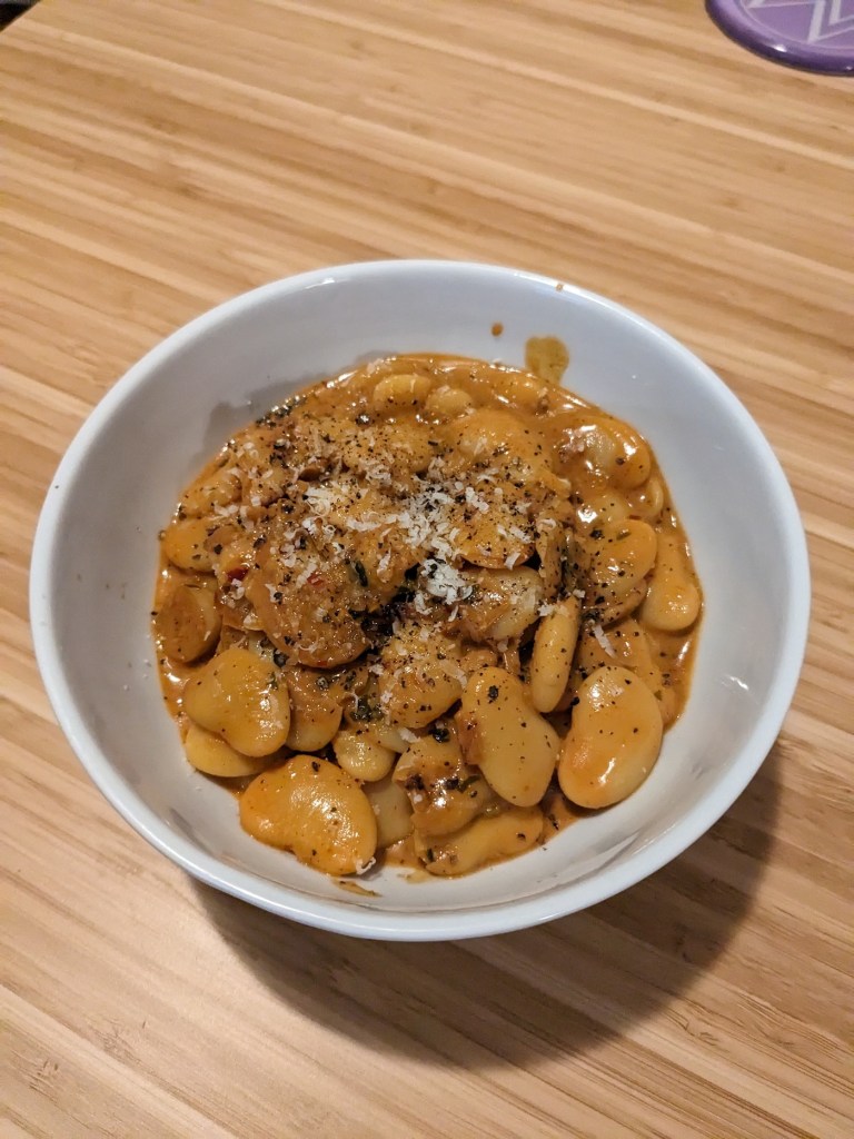 A bowl of big butter beans doused in an orange sauce. Grated parmesean and black pepper on top.