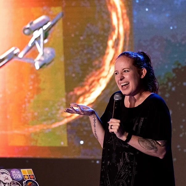an astrophysicist with really cool tattoos on her arm and with her hair tied in a ponytail smiles and talks into a mic with her hand propped out in gesture. behind her is a screen display of a futuristic space ship, from the star trek series, flying towards a planet