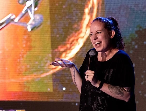 an astrophysicist with really cool tattoos on her arm and with her hair tied in a ponytail smiles and talks into a mic with her hand propped out in gesture. behind her is a screen display of a futuristic space ship, from the star trek series, flying towards a planet