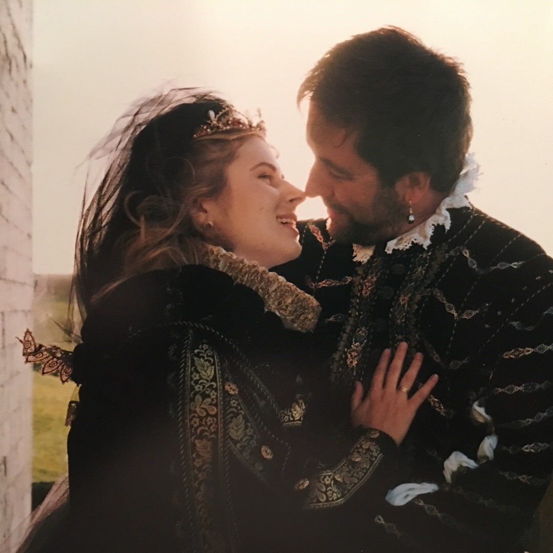 A man and a woman in Renaissance-period clothes looking into each others eyes as the sun sets behind them