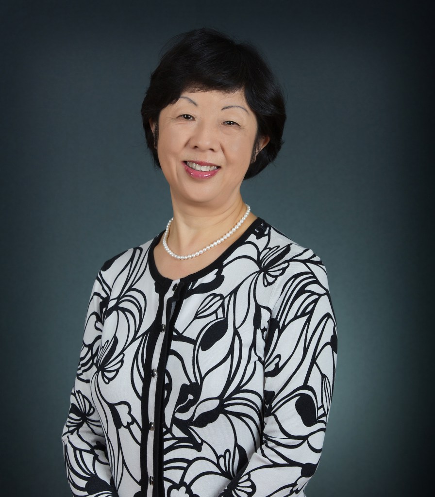 A woman (Xiaodong Lin-Siegler) standing and smiling at the camera wearing an elaborate patterned blazer.