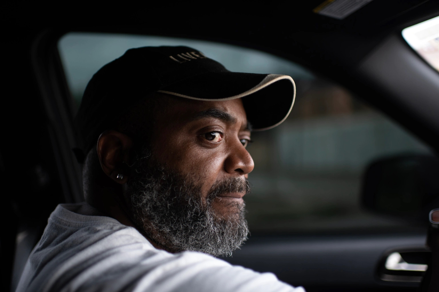 A man wearing a baseball cap drives a car near the oil refineries. 