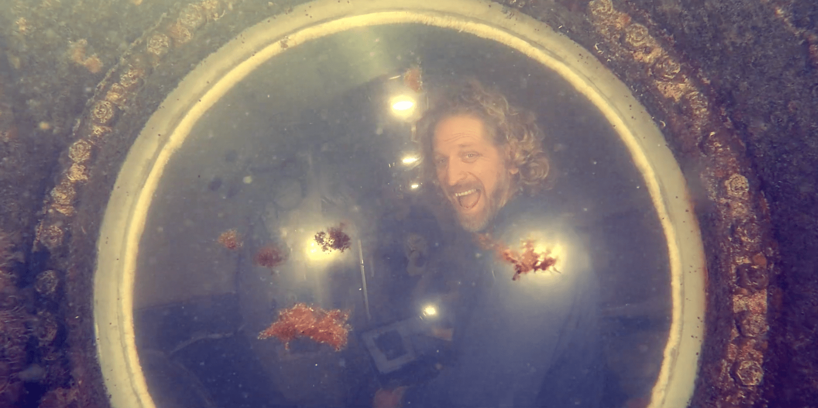 a man smiles through a circular window of an underwater lodge. the exterior of the lodge is slightly crusty with ocean debris and you can see floating debris in the water