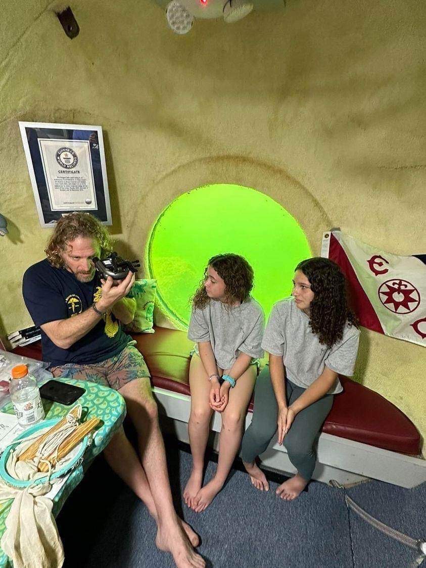 in a cavern like structure underwater, a man looks through a device on a couch. two teenagers sit next to him observing the undersea dwelling