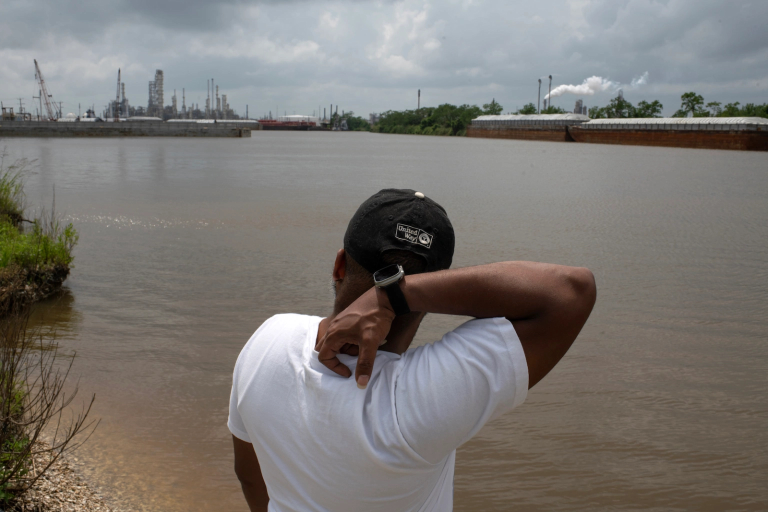 A man scratches his neck while looking out over a body of water to an oil refinery. 