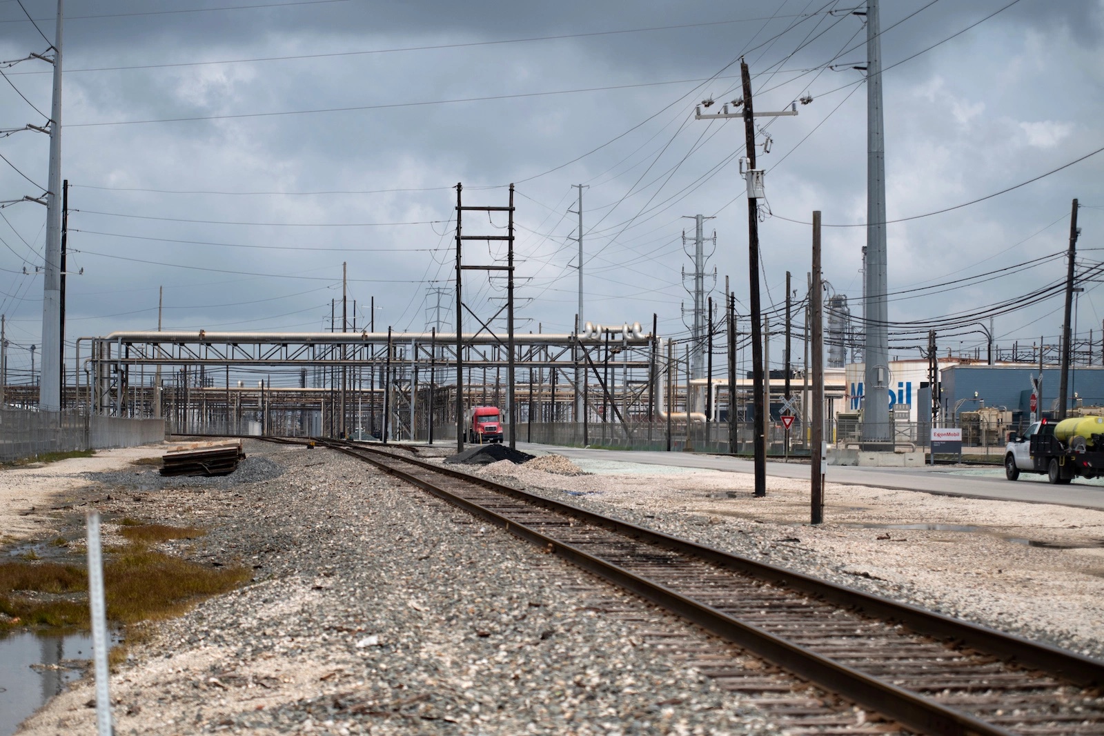 A red truck drives by a railroad track surrounded by industrial plants.