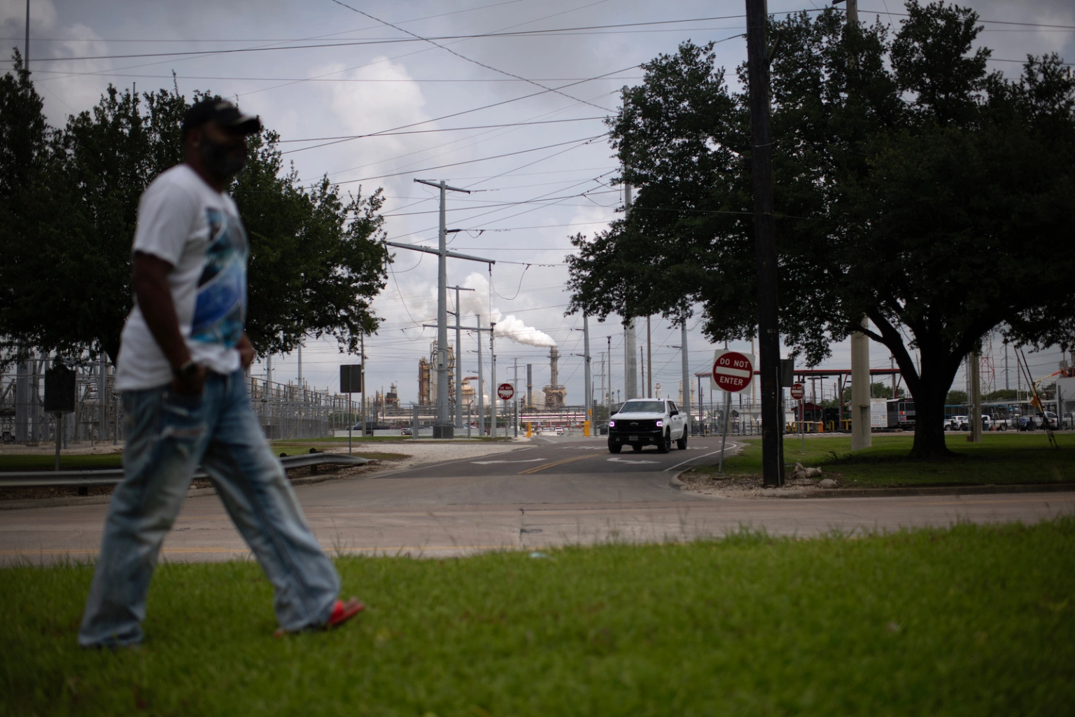 A man walks across a grassy yard, with a polluting oil refinery in the background. 