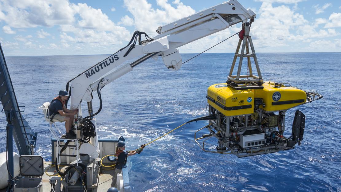 The large yellow robotic remotely operated vehicle is lifted by a hydraulic arm from the deck of of a ship and lowered into the bright blue ocean.