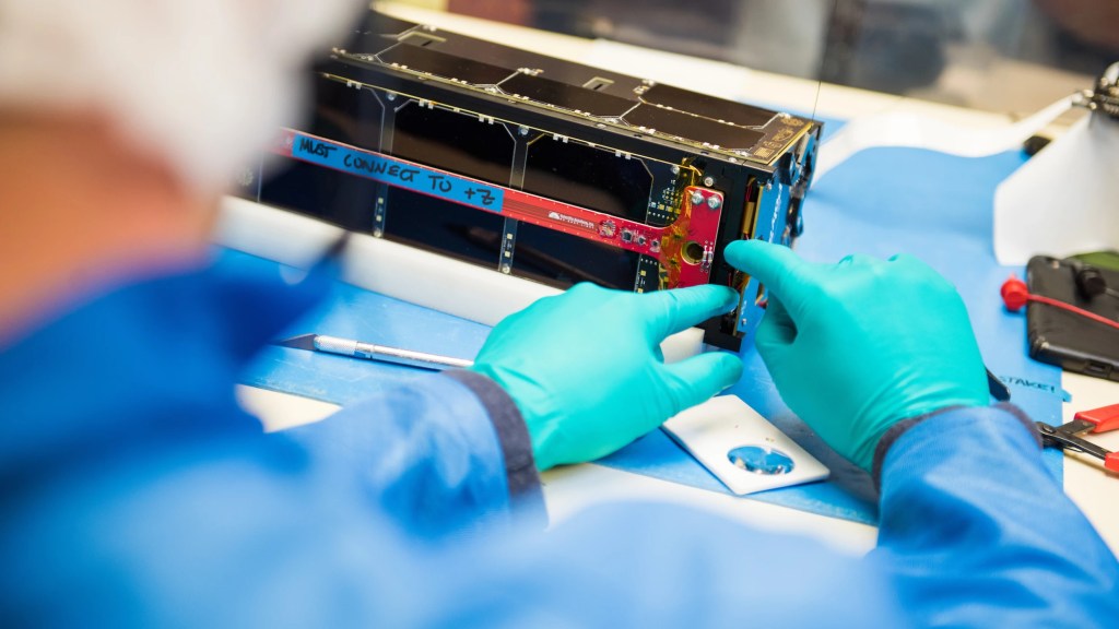 A person wearing gloves, tinkering with a shoe-box sized, half-finished boxy piece of technology. 