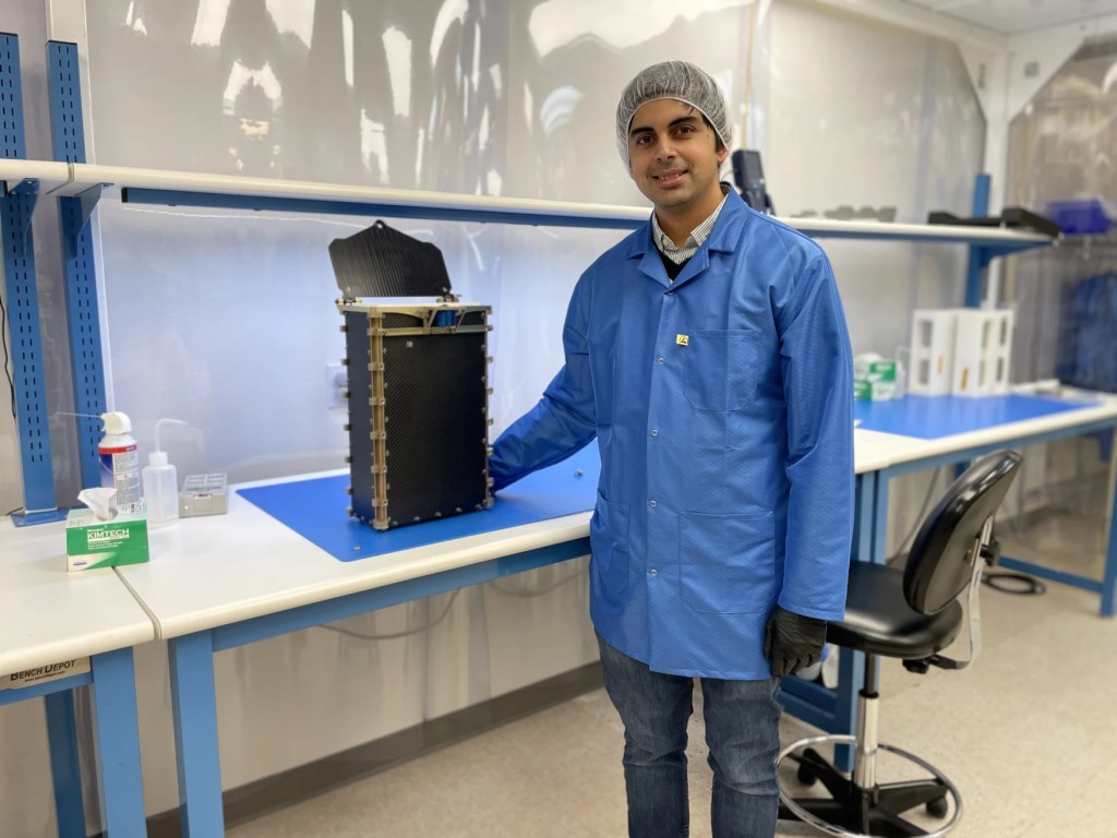 A man wearing a hairnet and a lab coat stands by a large CubeSat in a lab