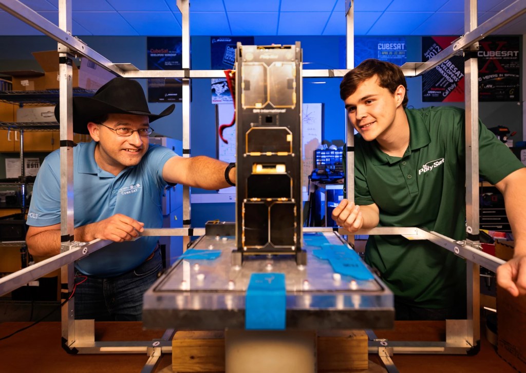 A man wearing a cowboy hat and younger man admire a larger CubeSat