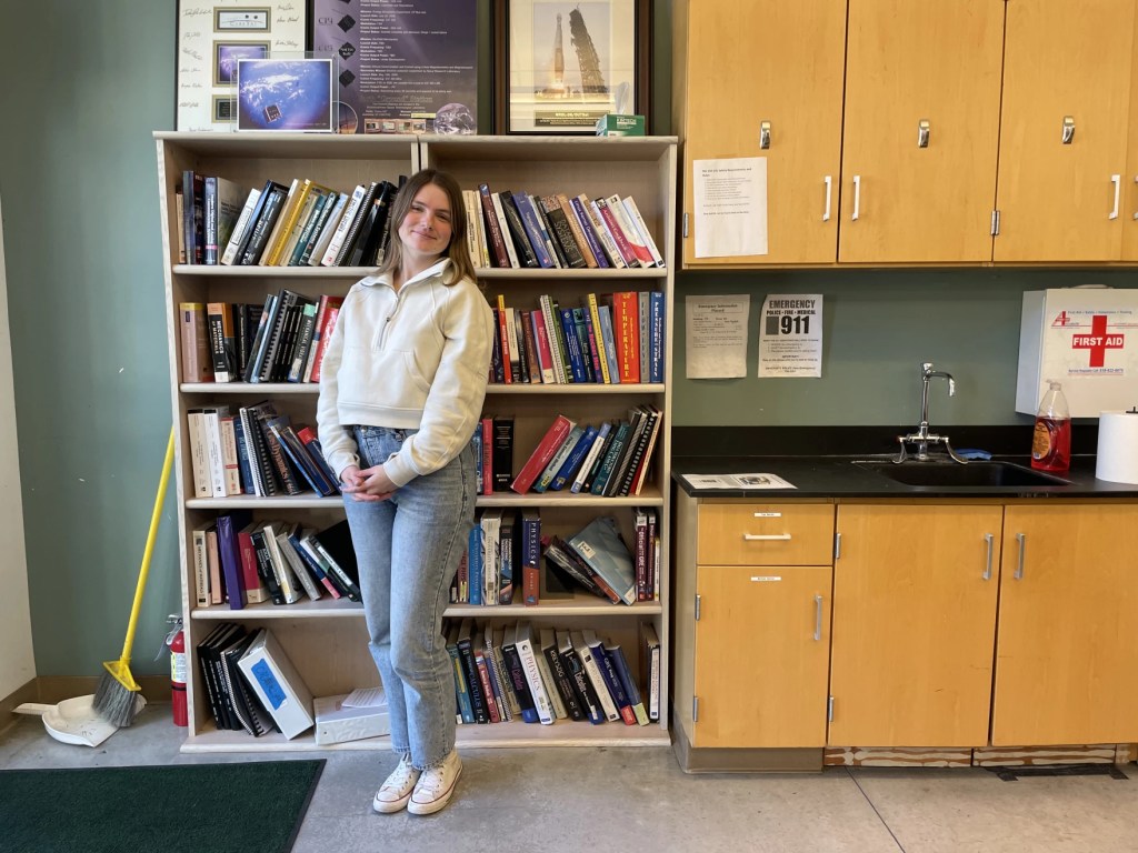 A young woman stands by a bookshelf in a lab.