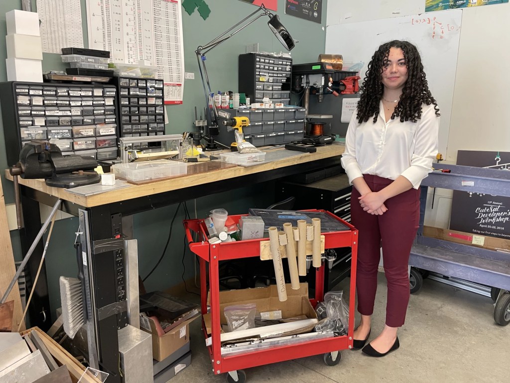 A young woman stands at a cluttered lab table full of half-finished engineering projects. 