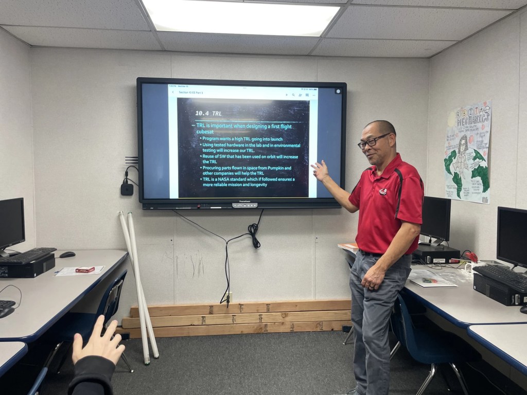 A man stands by a large monitor, presenting a slide to students