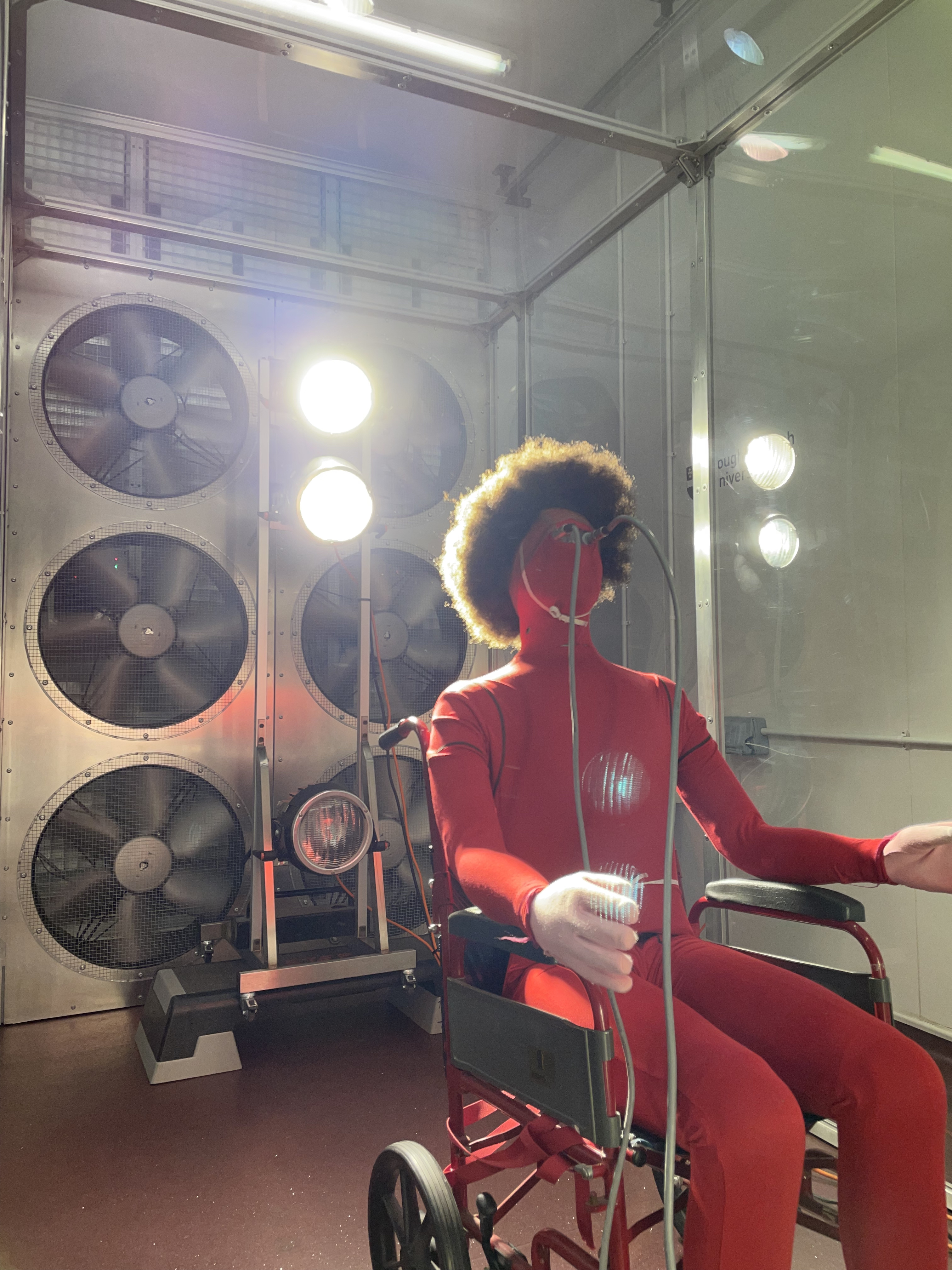 A manekin wearing all red and a curly afro wig sits in a wheelchair facing away from a wall of fans. Wires are plugged into its eye sockets. 