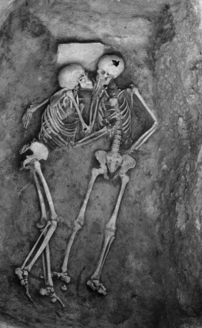 Two skeleton in an expedited grave facing one another. The hand of the skeleton on the left rests on the side of the skull of the skeleton on the right.