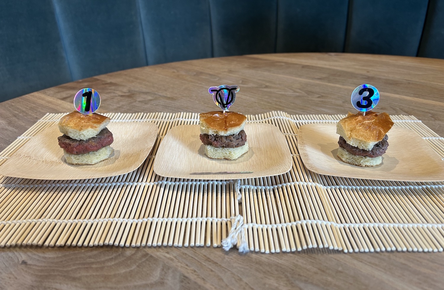 three small burgers each on separate plates that have different labels on toothpicks in the buns.