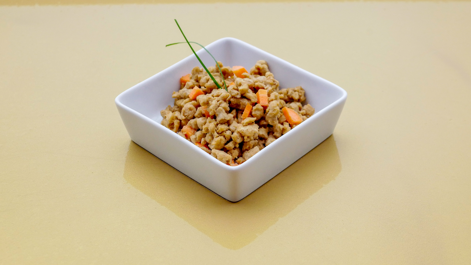 a square dish of brown cat food that has bits of carrots. a sprig of green onion sticks out from the chow