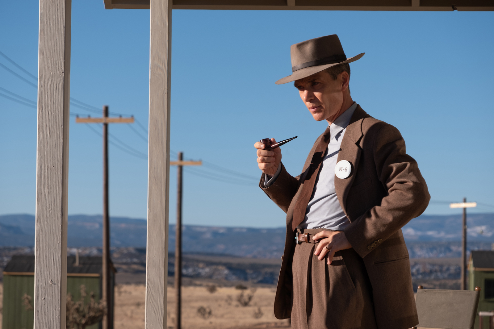 a man in a brown suit and brown fedora smoking a pipe in a desert like landscape.