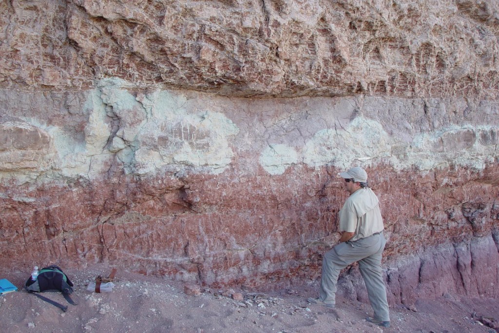 A man stands by a wall of colorful rock, arranged in stripes of brown, red, and beige.