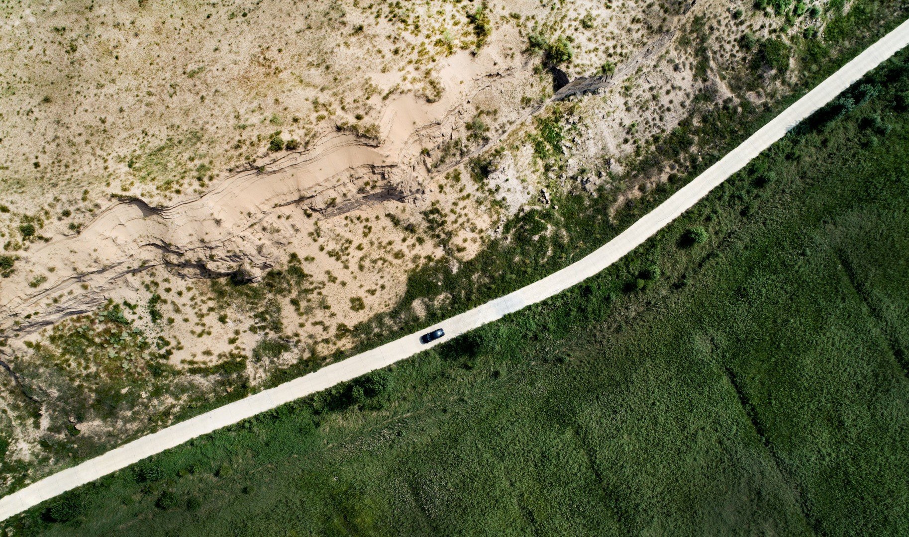 On one side of the road, there is a sandy desert with shrub-like green plants sparsely covering the soil. The other side of the road is lush with bright green with trees and plants. A black car drives along the bright white road.