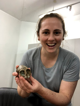 A woman holding a very small primate skull, smiling into the camera. 