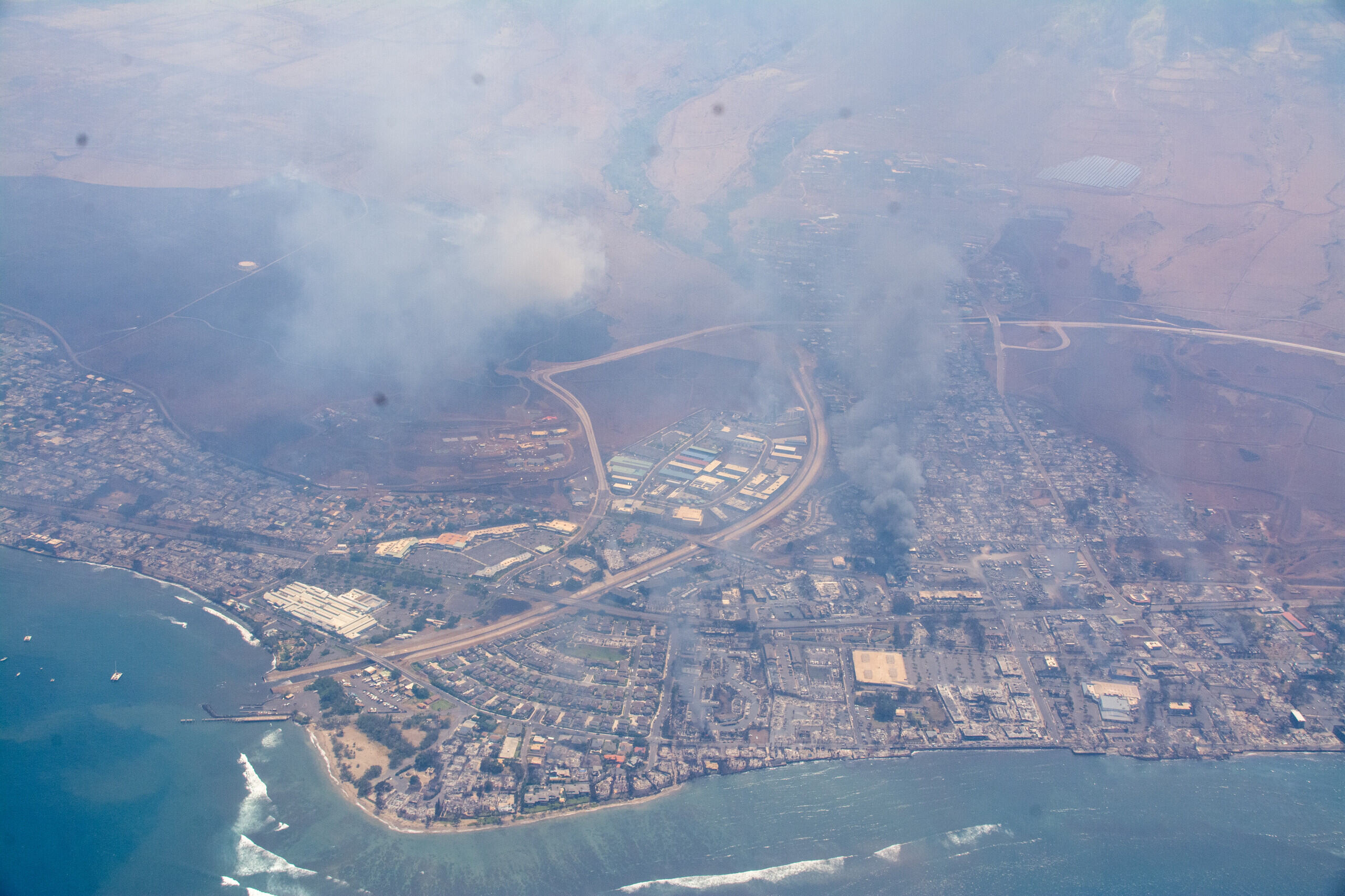 An aerial photo shows a ocean adjacent city in Maui burning with large plums of grey smoke.