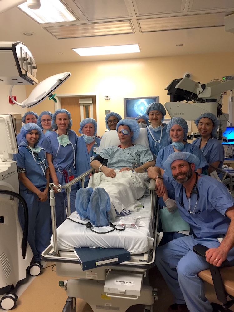 a group of health care workers in surgical gowns surround a patient with an eye patch who underwent surgery. they all smile at the camera
