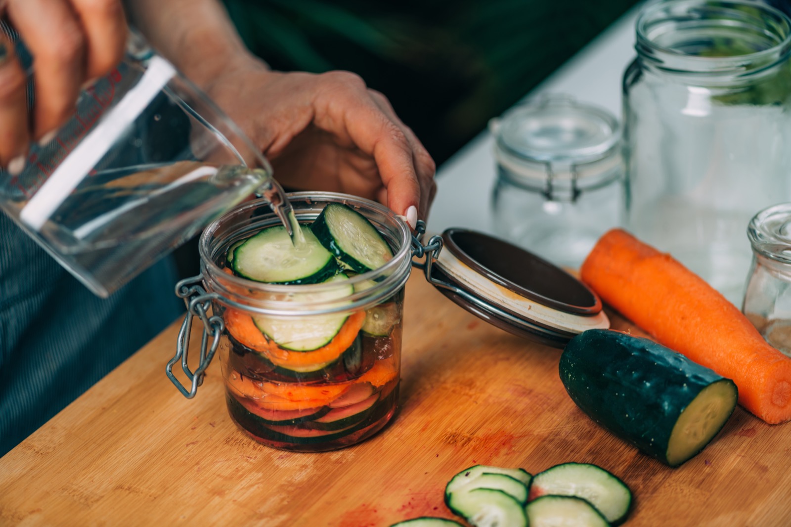 On a wooden cutting board a cucumber and carrot have been sliced. Slices of the vegetables have been placed in a glass canning jar. A person is pouring water over the vegetables in the jar.