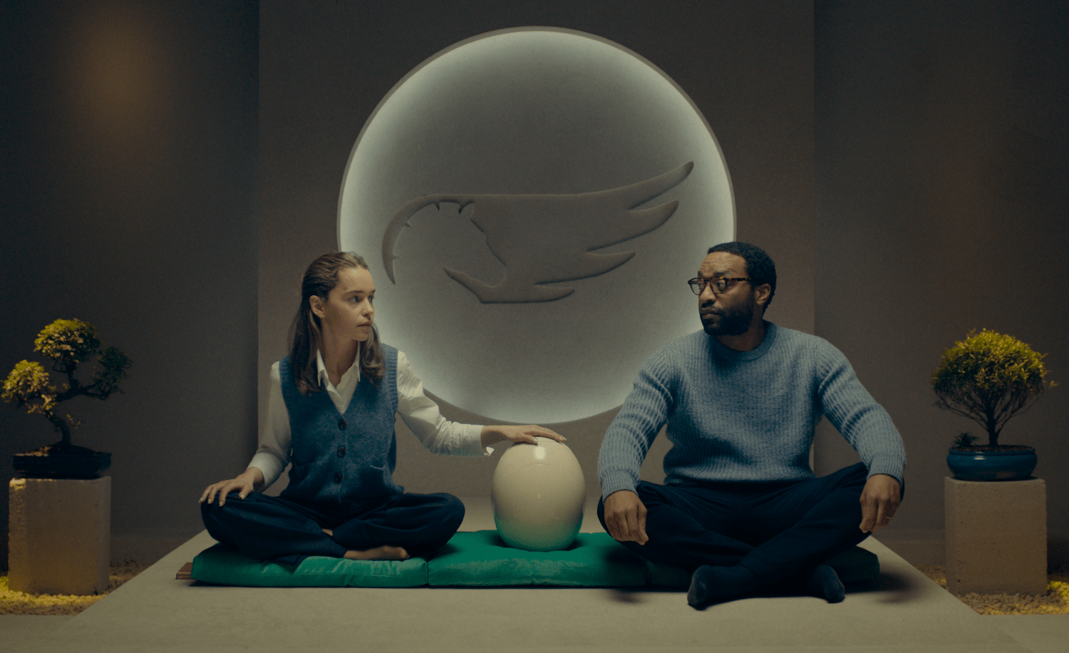 a black man and a white woman sit by a white egg shaped capsule. behind them is a logo for a horse with a wing