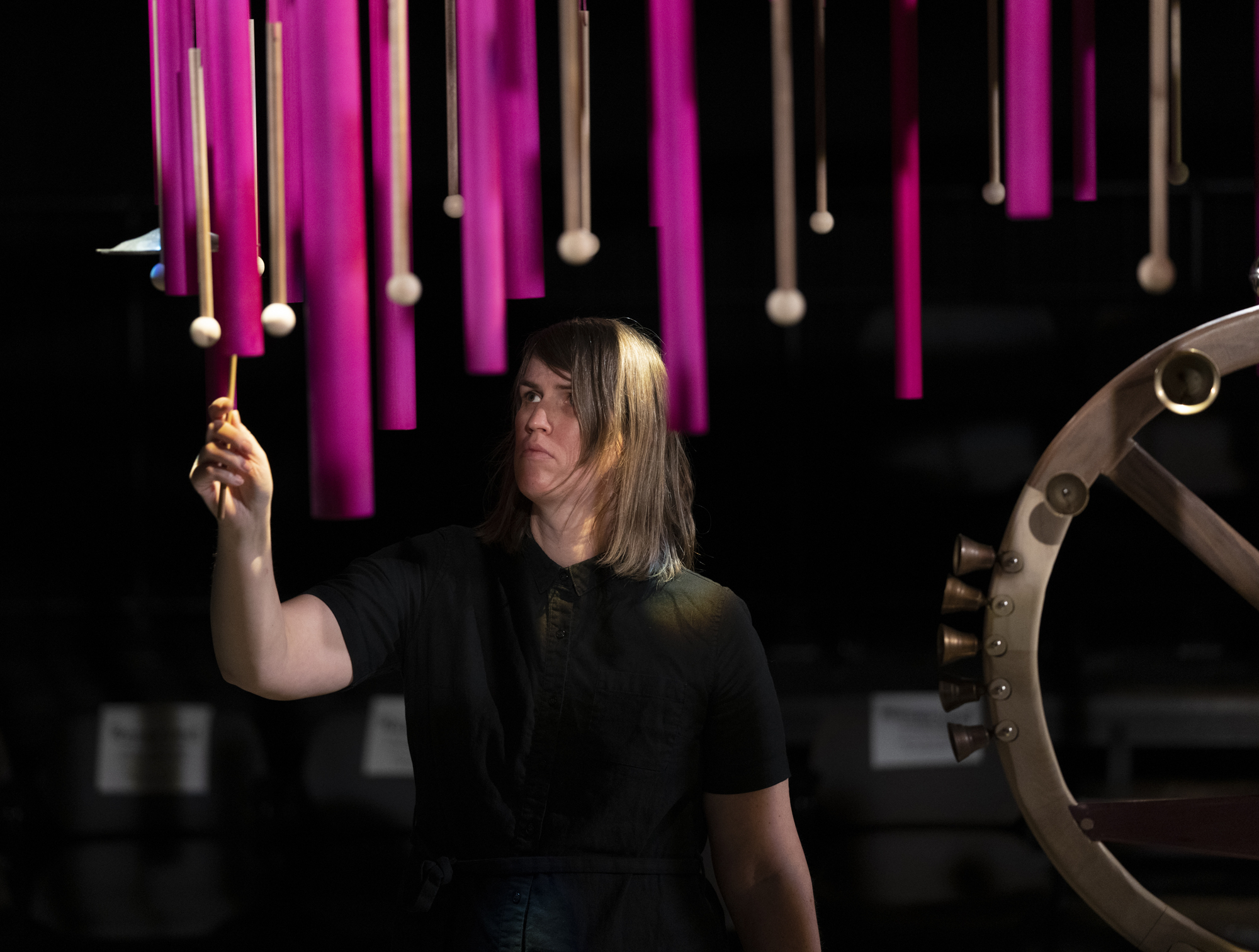 A white woman in a black shirt in a dark room raises a mallet among various brown mallets and hot pink pipes hanging down around her.