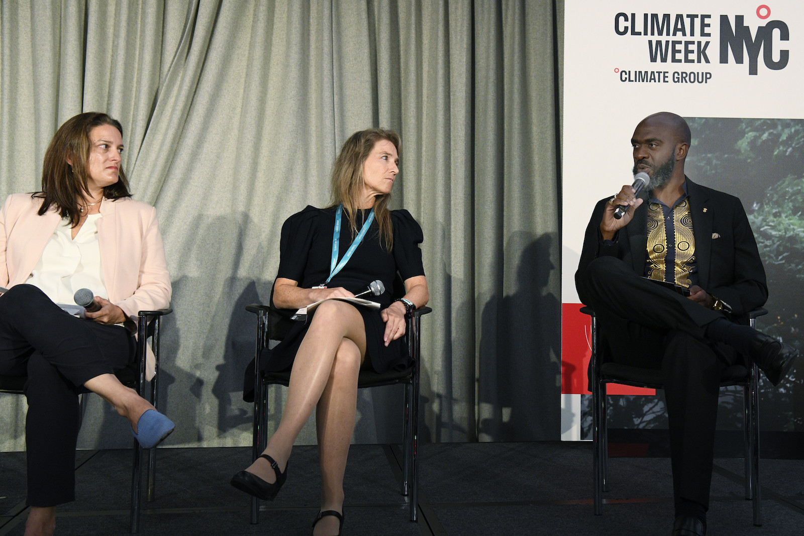 Three people sitting on a stage. A man on the right is holding a mic and behind him is a poster that says "NYC Climate Week"